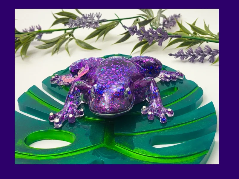 “Front-facing purple glitter resin frog with a butterfly on its back, sitting on a green acrylic monstera leaf.