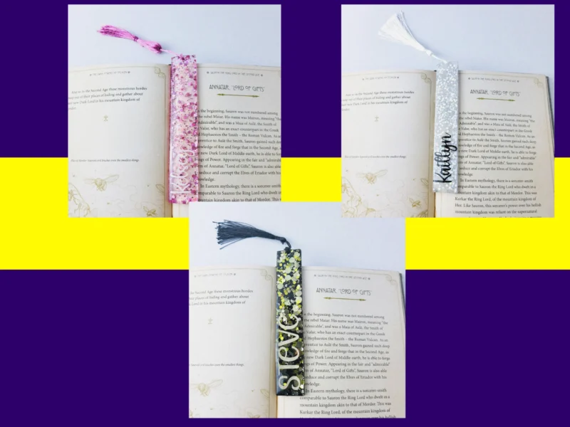 Three glitter resin bookmarks in pink, silver and black displayed inside an open fantasy book, each with matching tassels.