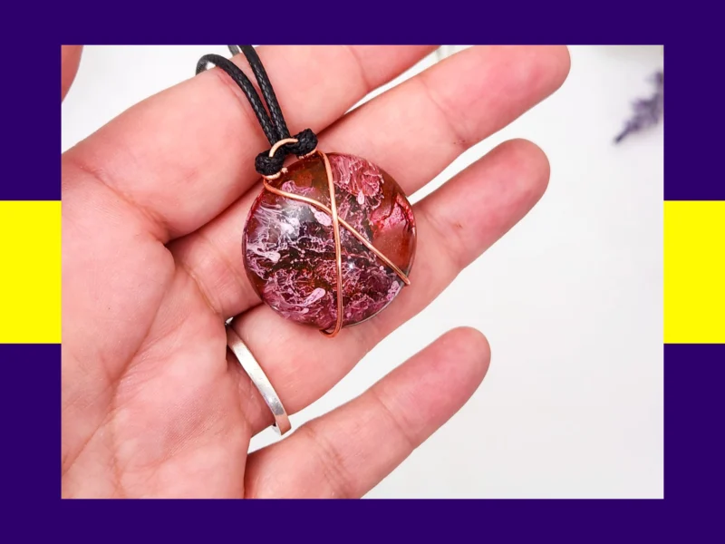 Round resin pendant in pink, burgundy and black with copper wire wrap shown in hand – handmade autumn inspired necklace