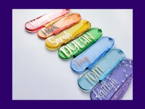 Oval glitter resin bookmarks in a rainbow of colours with personalised names and tassels arranged in a row.