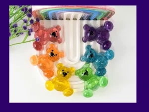 Six glitter resin Worry Bears in rainbow colours arranged on arch tray with purple flowers, colourful anxiety calming gifts