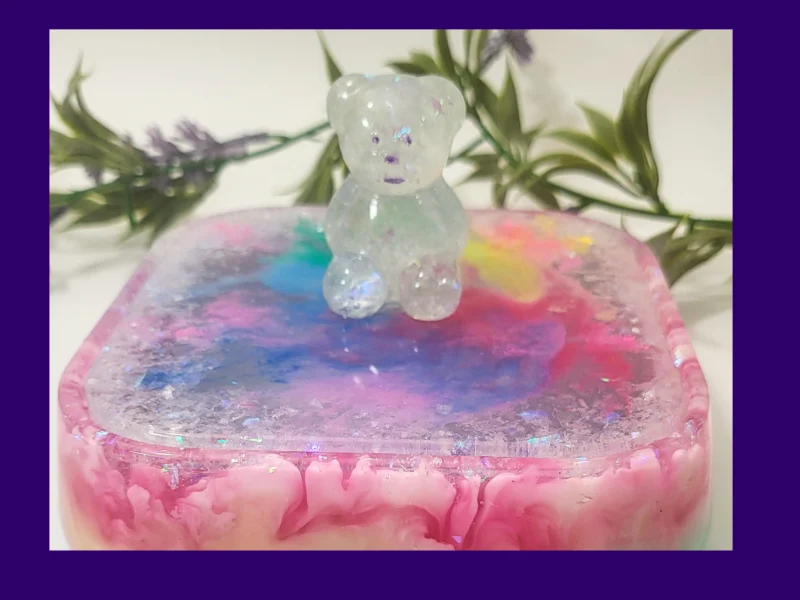 Close-up of a clear glitter teddy sitting on the lid of a pastel rainbow resin treasure box with iridescent sparkle flakes.