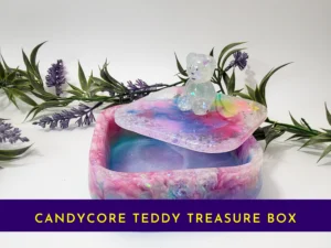 Open pastel rainbow resin treasure box with teddy-topped lid resting on the rim; banner text reads “Candycore Teddy Treasure Box”.