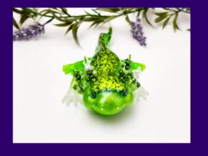 Front-facing glitter green resin axolotl surrounded by decorative foliage.