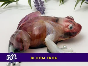 Side view of the Bloom Frog resin sculpture with a marbled burgundy/amber body and pearly legs; a purple banner along the bottom reads “BLOOM FROG” with a small KN logo.