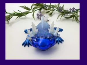 Fire & Ice axolotl (ice version) in blue glitter resin with translucent frills, photographed on a white background with greenery, framed by a purple border.