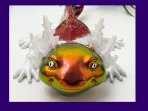 Fire axolotl with iridescent gold-green face and red glitter back on a white background with greenery, framed by a purple border.